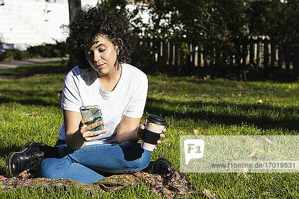 Young woman with disposable coffee cup using mobile phone while sitting at park