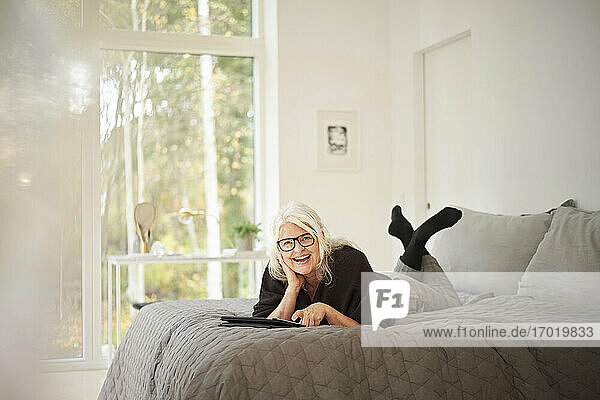 Happy senior woman with digital tablet lying on bed in bedroom