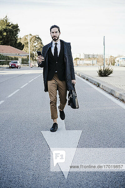 Businessman with briefcase and mobile phone walking on road