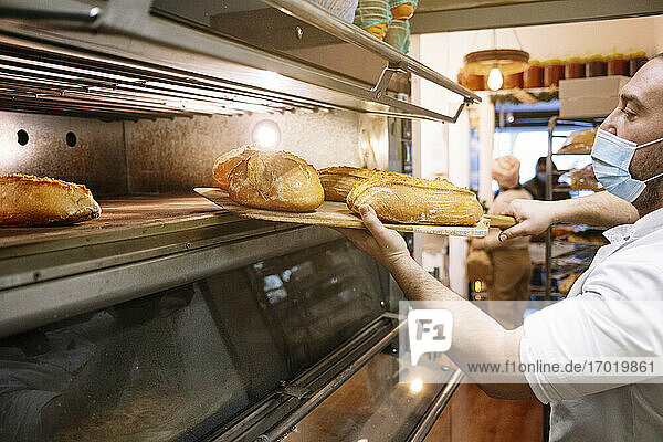 Männlicher Koch mit Gesichtsschutz  der in einer Bäckereiküche frisches Brot aus dem Ofen nimmt