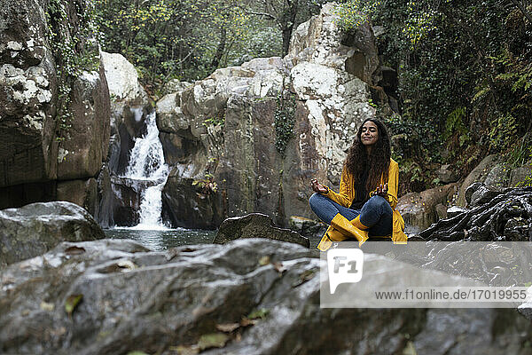 Frau in gelbem Regenmantel meditiert im Sitzen auf einem Felsen vor einem Wasserfall im Wald