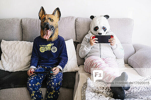 Geschwister in Tierkostümen spielen ein Handheld-Videospiel  während sie zu Hause auf dem Sofa sitzen