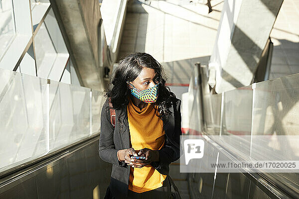Nachdenkliche Unternehmerin mit Mobiltelefon auf der Rolltreppe während der COVID-19
