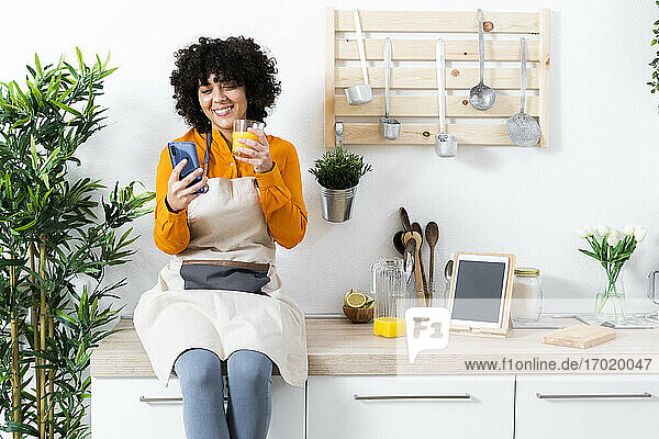 Lächelnde Frau  die ein Mobiltelefon benutzt und Orangensaft trinkt  während sie zu Hause auf dem Tisch sitzt