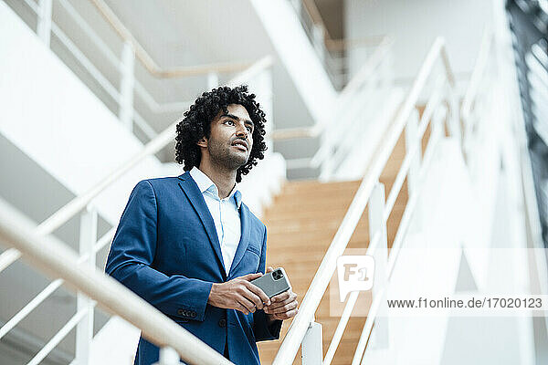 Nachdenklicher Geschäftsmann  der sein Smartphone hält und wegschaut  während er an der Treppe im Büro steht