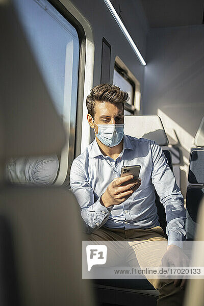Businessman with protective face mask using mobile phone while sitting in train during COVID-19