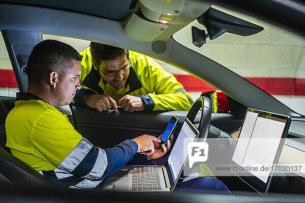 Male programmer showing mobile phone to colleague while sitting in electric car