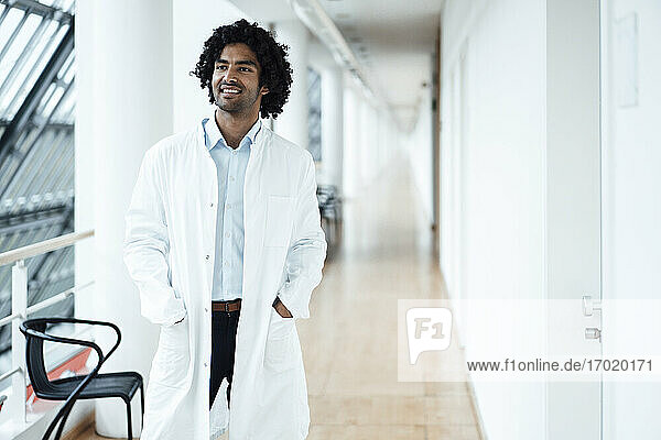 Lächelnder junger männlicher Arzt  der mit den Händen in den Taschen steht und auf den Krankenhausflur blickt