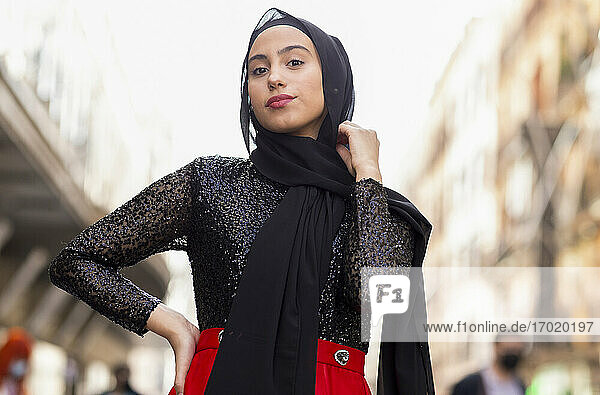 Portrait of young woman wearing black hijab posing outdoors with hand on hip