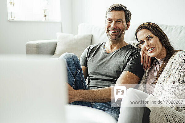 Lächelndes reifes Paar  das einen Film auf dem Laptop ansieht  während es im Wohnzimmer zu Hause sitzt