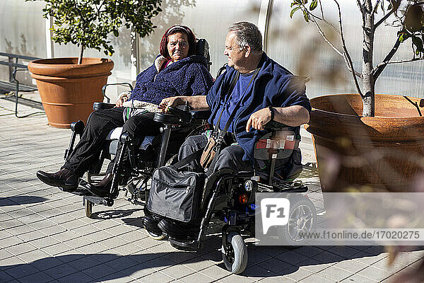 Behinderte Frau und Mann halten sich an den Händen  während sie an einem sonnigen Tag im Rollstuhl sitzen