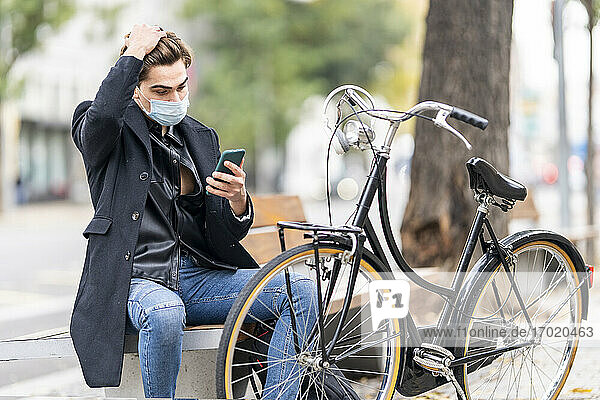 Junger Mann mit Hand in den Haaren  der sein Smartphone benutzt  während er auf einer Fahrradbank in der Stadt sitzt  während COVID-19