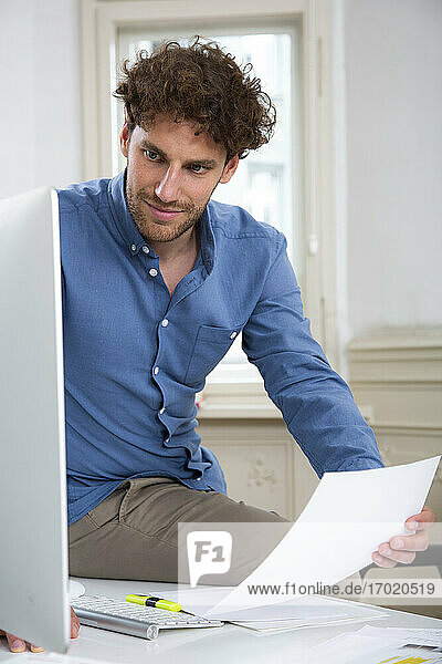 Junger Geschäftsmann arbeitet am Computer mit einem Dokument am Schreibtisch im Büro