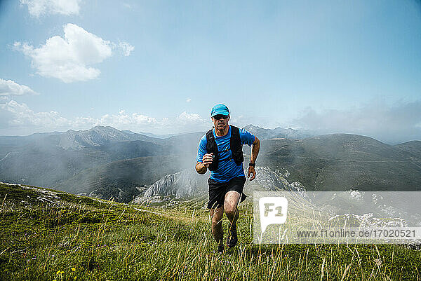 Älterer Sportler beim Trailrunning in den Bergen auf einer Wiese vor blauem Himmel