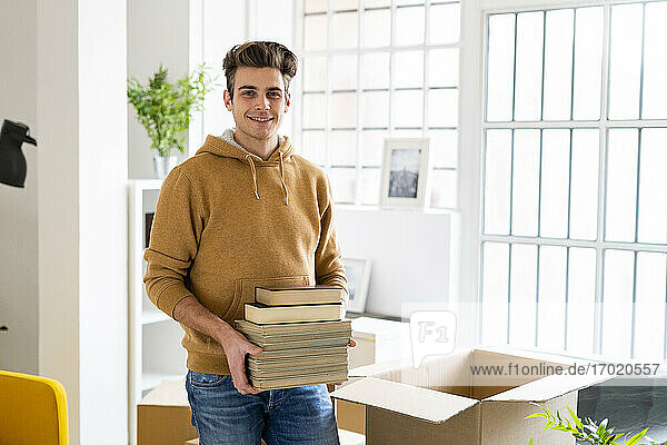 Lächelnder junger Mann mit Büchern in einem Karton in seiner neuen Loftwohnung