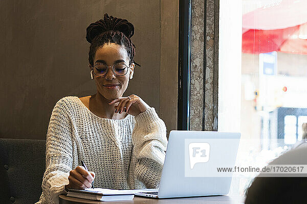Lächelnde junge Frau  die mit einem Laptop in einem Café sitzt und in ihr Tagebuch schreibt
