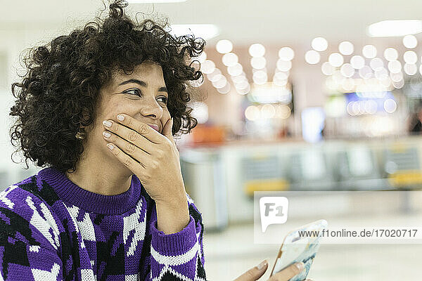 Young woman laughing while using mobile phone sitting at airport