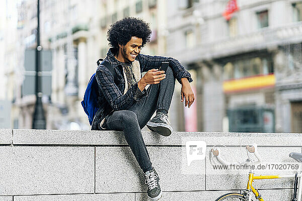 Smiling man with curly hair using mobile phone while sitting on retaining wall in city