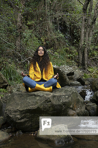 Female hiker wearing yellow raincoat meditating while sitting on rock in forest