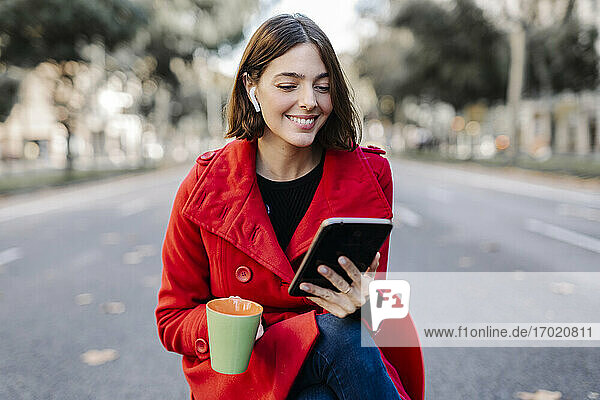 Smiling fashionable young woman with coffee cup using digital tablet on street