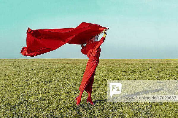 Senior woman holding red fabric while standing on grass against blue sky