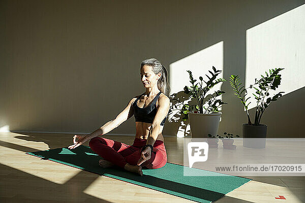 Yogini meditiert auf einer Übungsmatte im Wohnzimmer an einem sonnigen Tag