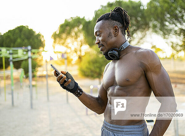 Sportler mit Kopfhörern  der im Park stehend ein Mobiltelefon benutzt