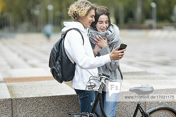 Junge Freunde mit Fahrrad lachen  während sie auf dem Gehweg ein Video über ihr Smartphone ansehen