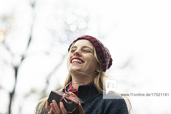 Fröhliche junge Frau mit Strickmütze  die ein Geschenk gegen den klaren Himmel in der Weihnachtszeit hält