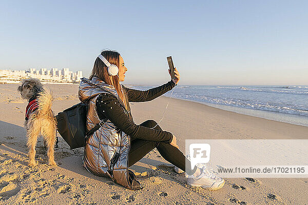 Woman taking selfie while sitting with dog at beach against clear sky during sunset