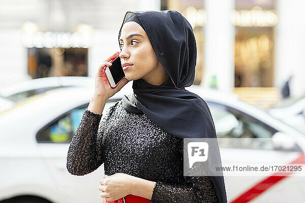 Portrait of young beautiful woman wearing black hijab talking on smart phone in middle of street