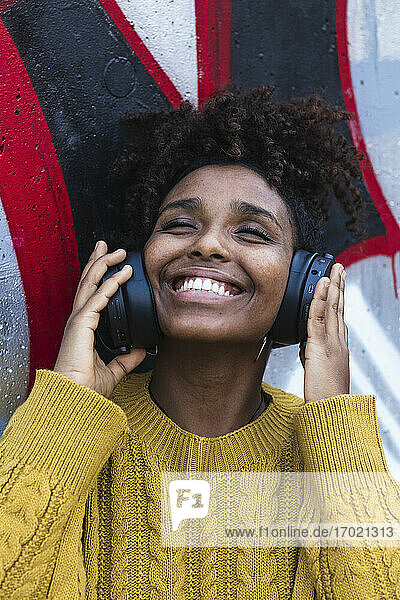 Glückliche junge Frau mit geschlossenen Augen  die über Kopfhörer an einer bemalten Wand Musik hört
