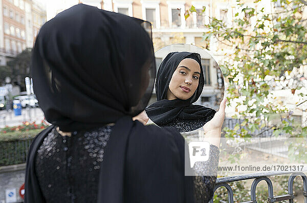 Junge schöne Frau mit schwarzem Hidschab steht im Freien und schaut in einen runden Spiegel