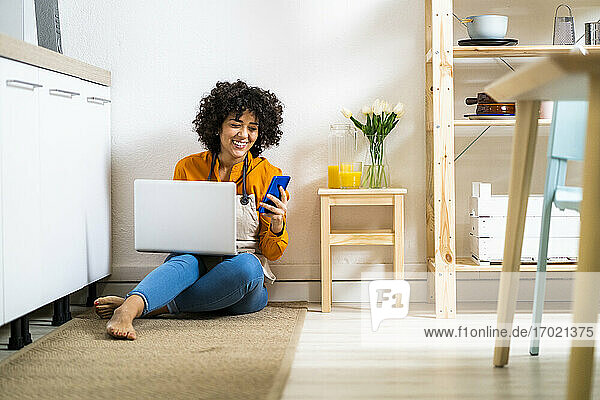 Junge Frau mit Laptop und Mobiltelefon  während sie zu Hause auf dem Boden sitzt