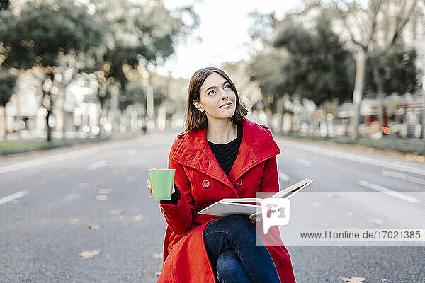 Junge Frau in roter Jacke mit Kaffeetasse träumt  während sie ein Buch auf der Straße hält