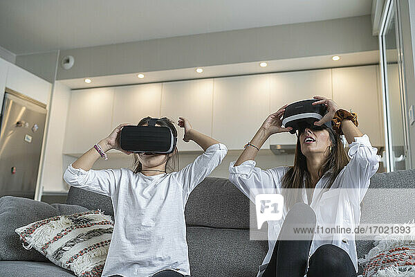 Mother using virtual reality glasses with her daughter while sitting on sofa in living room