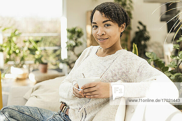Lächelnde junge Frau hält eine Kaffeetasse  während sie zu Hause auf dem Sofa sitzt
