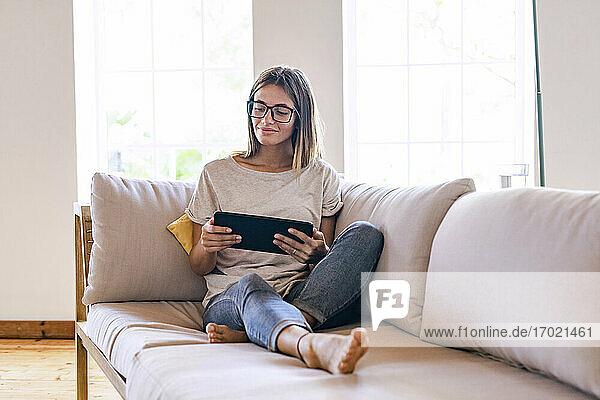 Entspannte brünette Geschäftsfrau, die auf dem Sofa sitzt und ein digitales Tablet im Wohnzimmer zu Hause benutzt