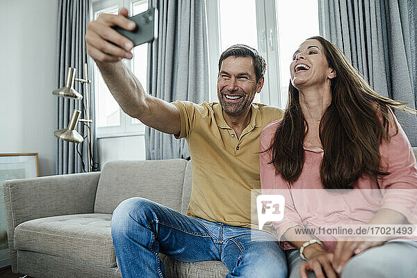 Älteres Paar lacht und nimmt Selfie beim Sitzen auf Sofa im Wohnzimmer
