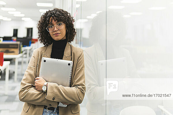 Eine Unternehmerin hält einen Laptop in der Hand  während sie im Büro im Flur vor einem Glas steht