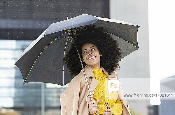 Lächelnde Frau mit Jacke und Regenschirm in der Hand im Regen stehend