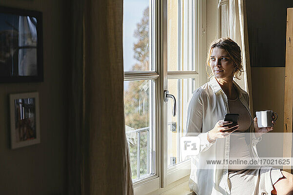 Junge schwangere Frau mit Kaffeetasse und Handy in der Hand  während sie durch das Fenster ihres Hauses schaut