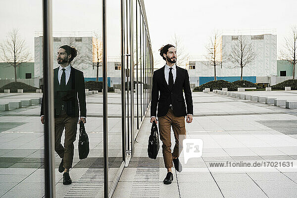 Young businessman with briefcase looking away while walking by glass wall