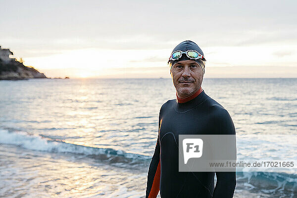 Portrait of male swimmer standing alone on coastal beach at sunset