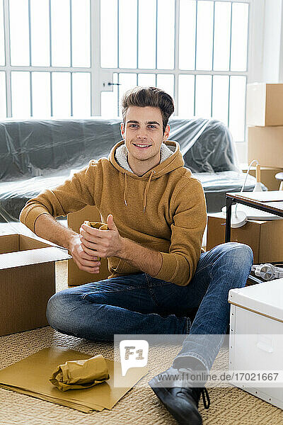 Lächelnder Mann  der ein eingewickeltes Trinkglas in einer Schachtel in seiner neuen Loftwohnung verpackt
