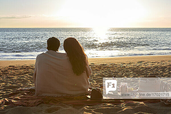 Junges Paar in Decke gehüllt am Strand sitzend und die Aussicht betrachtend