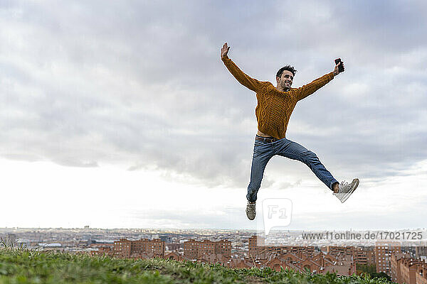 Excited young man holding mobile phone while jumping on hill against cityscape