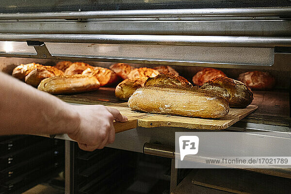 Männlicher Koch  der gebackene Brote mit einem Pizzaschieber aus dem Ofen in einer Bäckerei nimmt