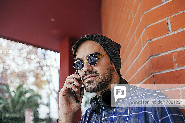 Modischer junger Mann mit Sonnenbrille beim Telefonieren gegen eine Backsteinmauer