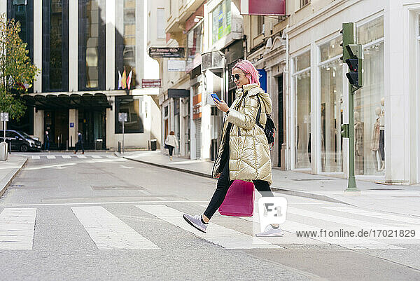 Junge Frau mit Einkaufstasche  die ein Mobiltelefon benutzt  während sie auf der Straße geht